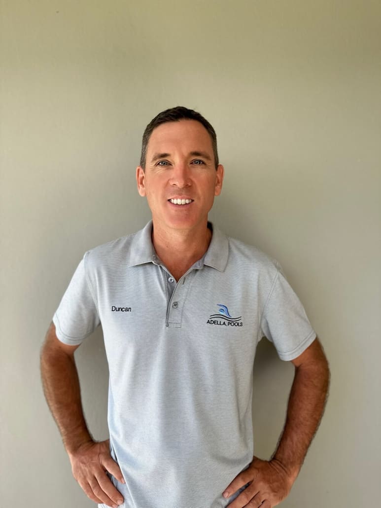 A man Duncan with short dark hair stands against a plain background, smiling at the camera. He is wearing a light grey polo shirt with the name "Duncan" embroidered on the left side and a logo on the right that reads "ADELLA POOLS" with a blue wave design. As one of Brisbane's top pool builders, his hands are confidently on his hips.