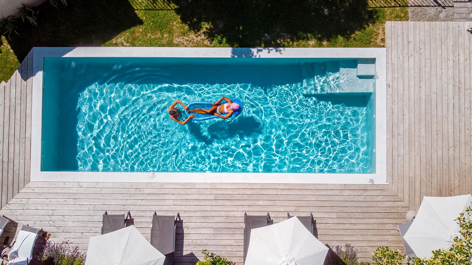 Aerial view of a rectangular outdoor pool with clear blue water, expertly crafted by Pool Builders Brisbane. Two people float on their backs in the center of the pool. Surrounding it is a wooden deck with several lounge chairs and umbrellas. Shadows from nearby trees partially cover the scene.