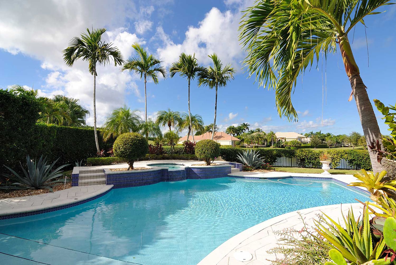 A clear blue swimming pool, expertly crafted by Pool Builders Brisbane, is surrounded by a well-maintained patio with various tropical plants and trees, including palm trees. The sky is bright and mostly clear, with a few scattered clouds. Beyond the pool area, you can see a fence and houses in the background.