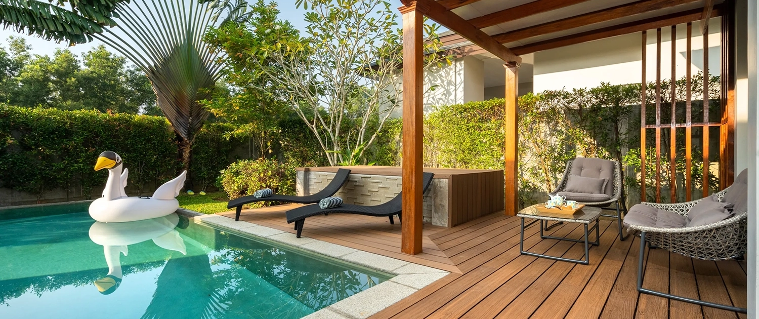 A backyard scene crafted by top Pool Builders Brisbane features a swimming pool with a large inflatable swan, two black lounge chairs, a hot tub, and a wooden deck with a pergola. On the deck are cozy chairs and a small glass-top table. Lush greenery surrounds the area, creating a tranquil escape.