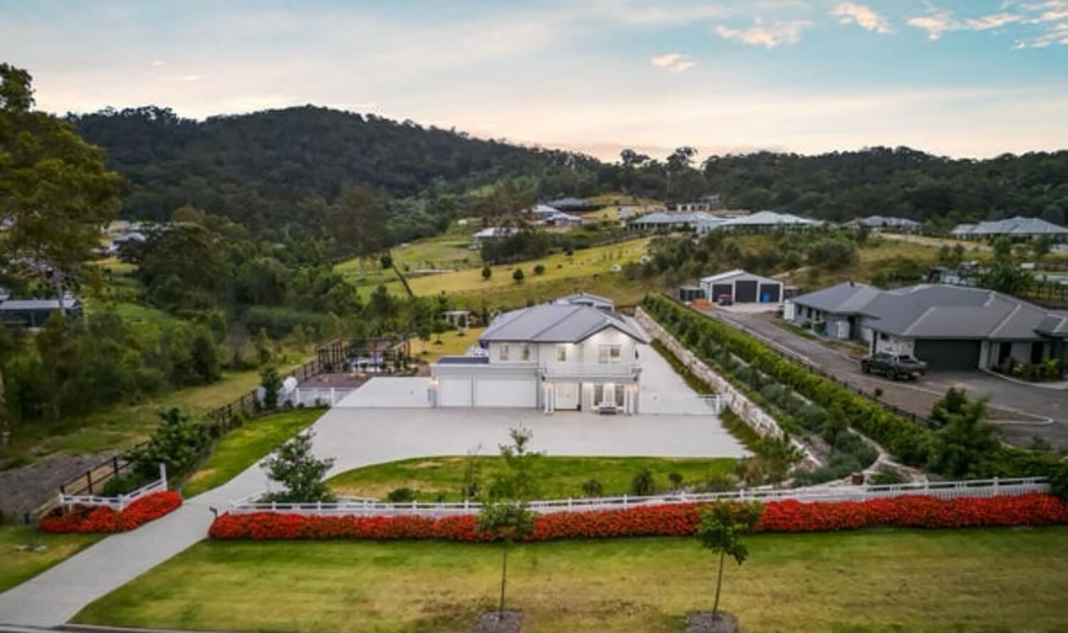 A luxurious two-story house with a spacious driveway, surrounded by a well-manicured lawn and vivid red flowers. The property is nestled in a picturesque, hilly landscape where Pool Builders Brisbane have crafted stunning pools for several nearby homes, set against the backdrop of a partly cloudy sky.