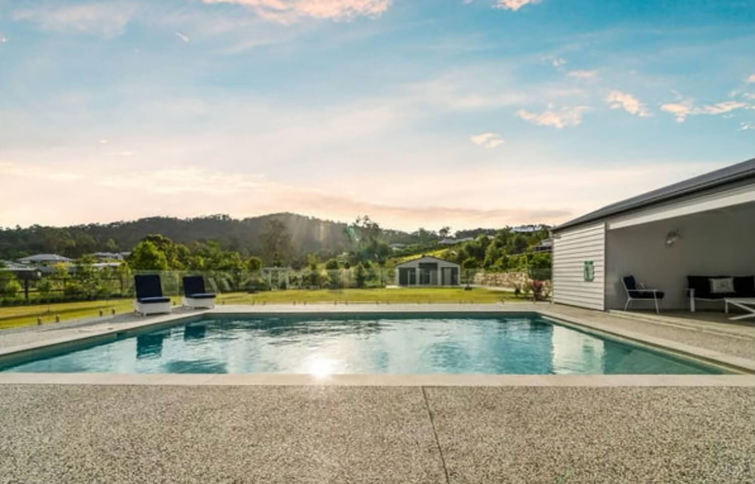 A serene outdoor scene featuring a large, rectangular swimming pool crafted by expert Pool Builders Brisbane, surrounded by a stone deck. Poolside lounge chairs are positioned for relaxation. In the background, there are rolling green hills, a small house, and a partly cloudy sky with the sun shining brightly.