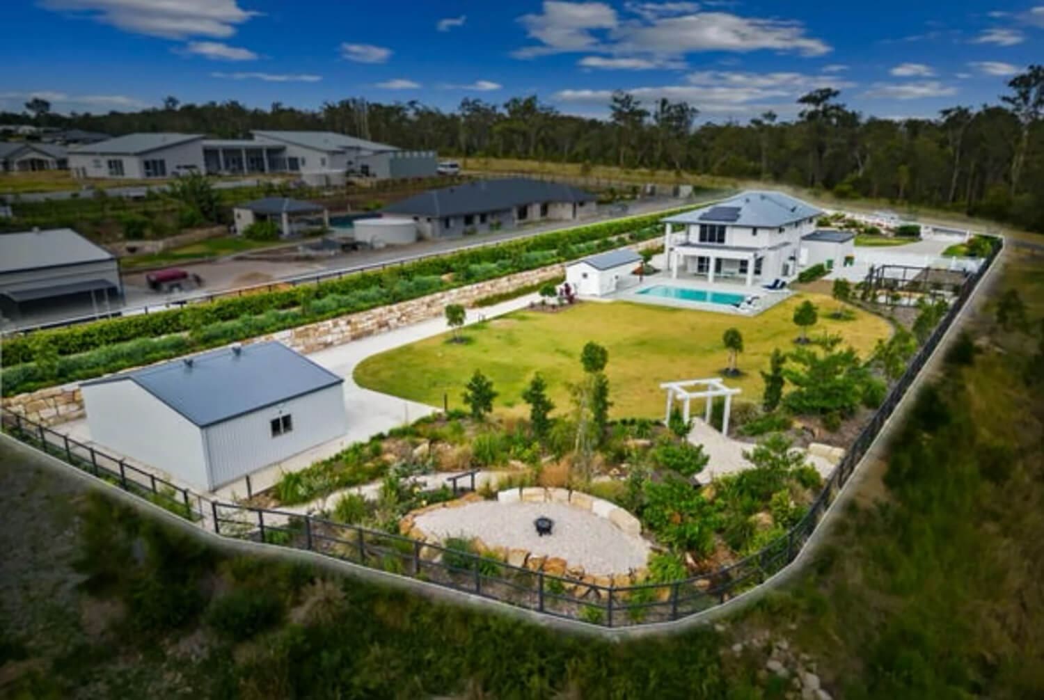 Aerial view of a large residential property featuring a modern house with a swimming pool installed by Pool Builders Brisbane, surrounded by well-maintained lawns, garden, and shrubbery. The property is enclosed by a fence and includes a circular garden area with a fire pit and a separate outbuilding.