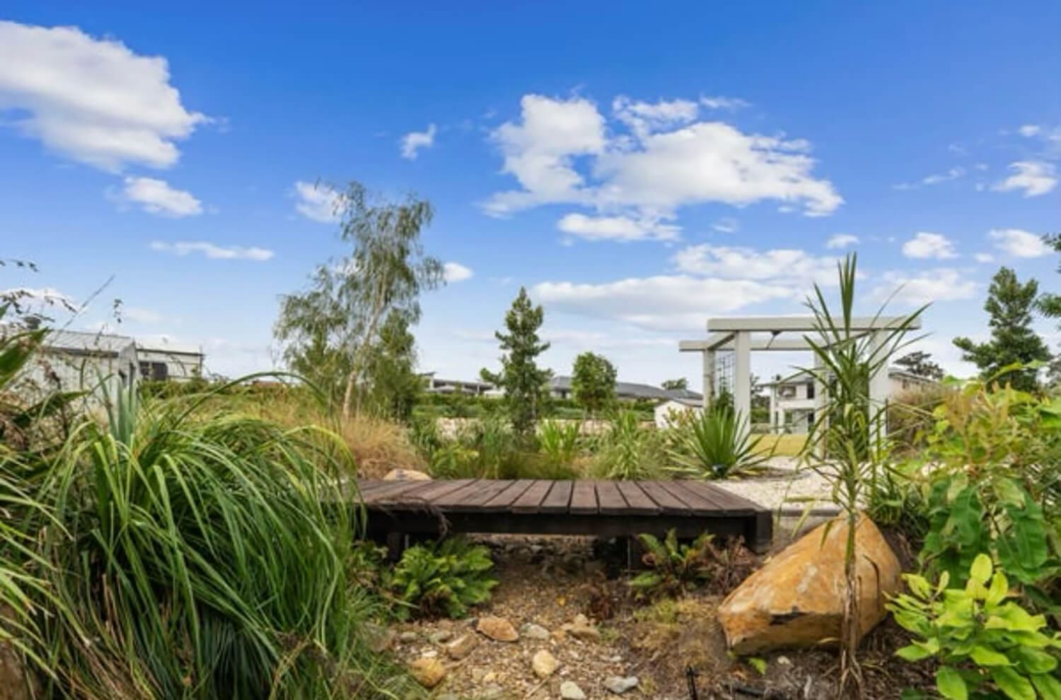 A serene garden landscape, meticulously crafted by Pool Builders Brisbane, features a small wooden bridge over a dry stream bed, surrounded by various lush green plants and large rocks. Trees and a white arbor are visible in the background under a bright blue sky with scattered clouds.