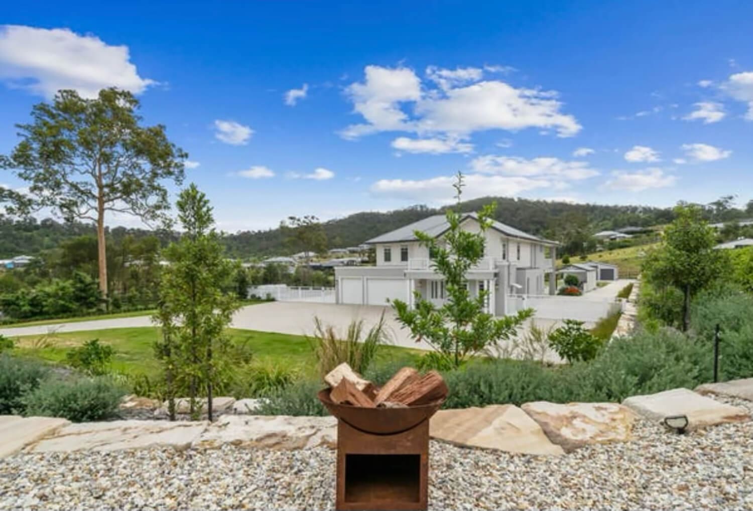 A modern two-story white house with large windows and a spacious yard is set against a backdrop of green hills and a blue sky with scattered clouds. In the foreground, expertly crafted by Pool Builders Brisbane, a metal fire pit with logs sits on a pebble-covered area surrounded by various plants.