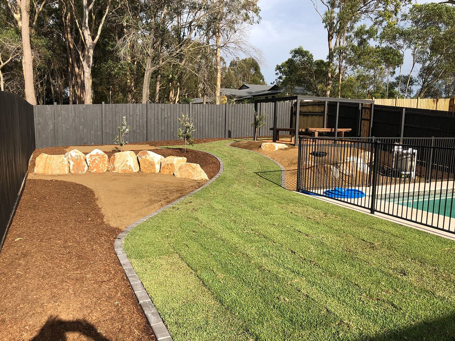 A backyard features a manicured lawn, a fenced swimming pool by Pool Builders Brisbane, and a patio area with seating under a pavilion. There are garden beds with large rocks and mulch near black fencing that encloses the backyard. Trees and foliage are visible in the background.