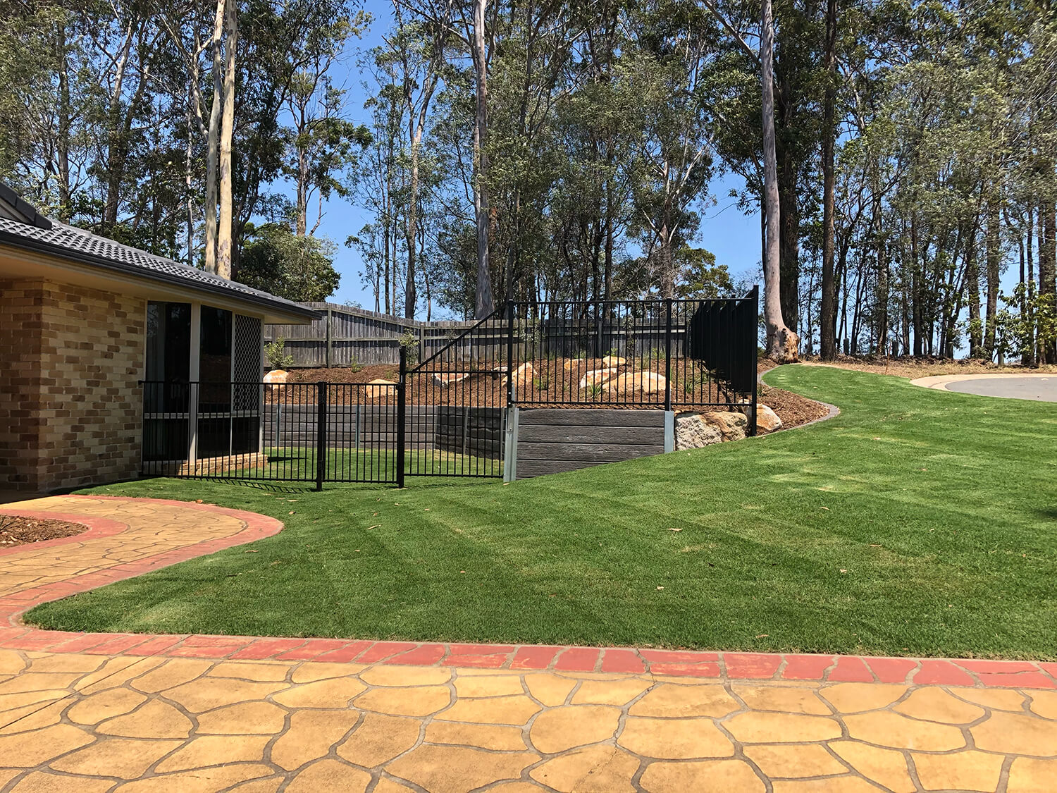 A backyard with a well-maintained lawn, stone walkway, and black metal fence. To the left is a brick house with large windows. In the background, there are tall trees and a sunny, clear sky. The scene is peaceful and picturesque—perfect for envisioning your dream pool built by Pool Builders Brisbane.