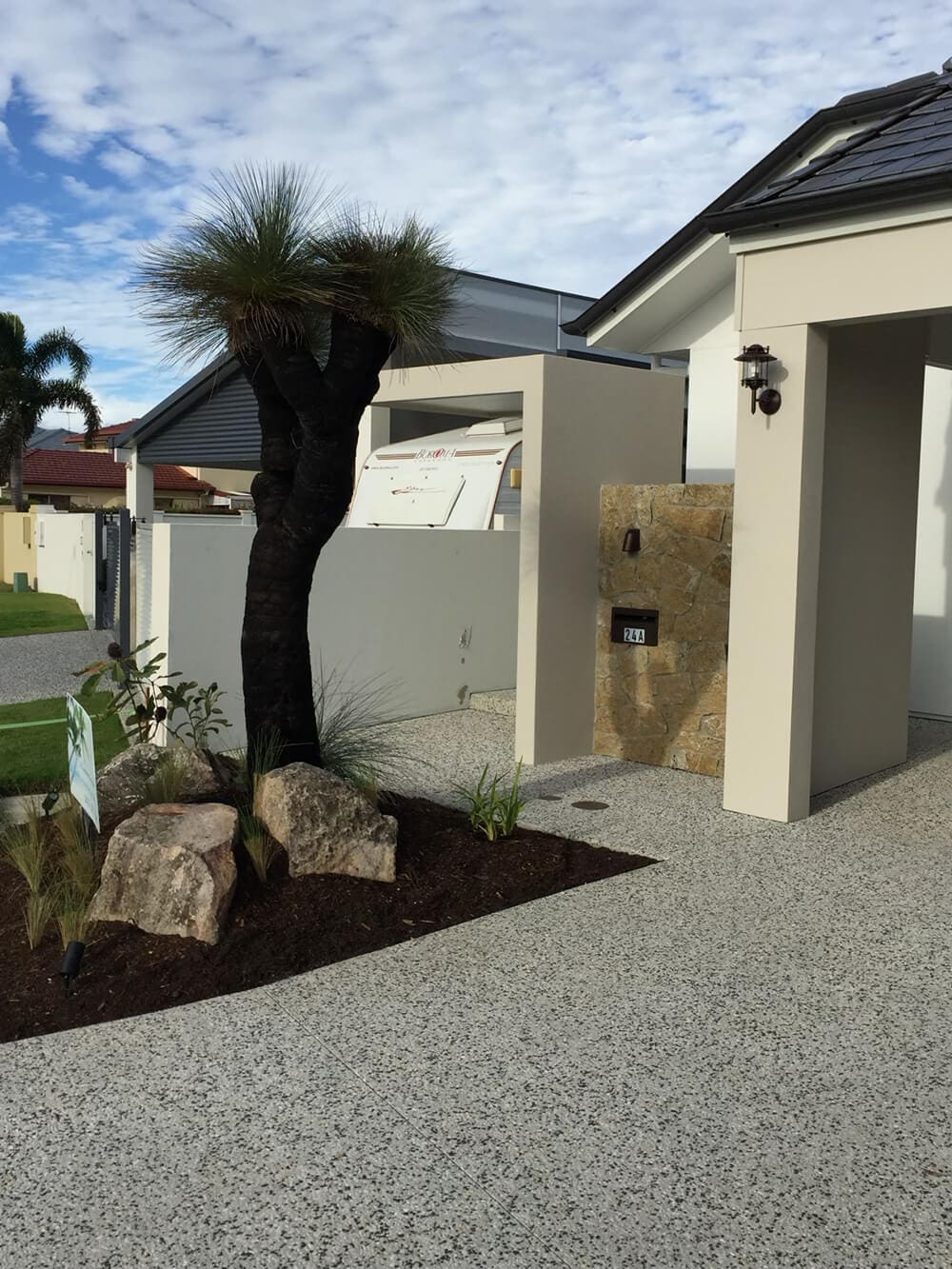 Modern house exterior with a small landscaped front yard featuring a black tree, rocks, and plants. The entrance has a beige stone wall with a house number "120" and a light fixture. A project admired by Pool Builders Brisbane, the sky is partly cloudy.