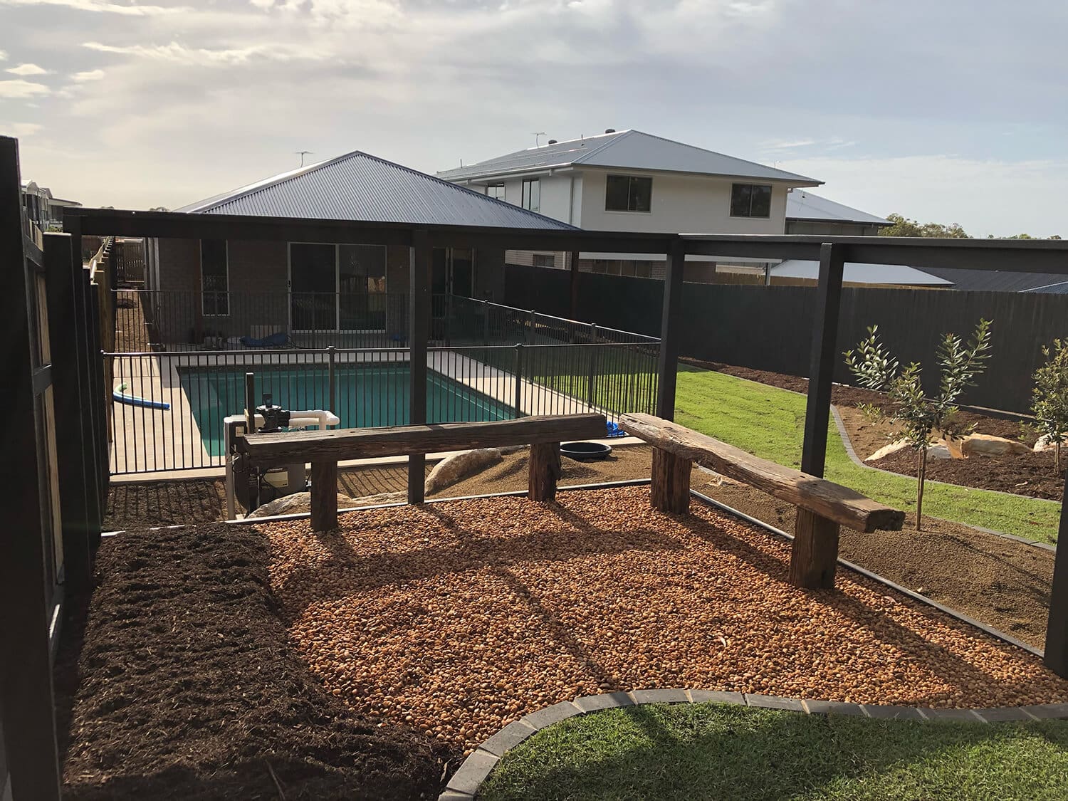 A backyard scene crafted by skilled Pool Builders Brisbane showcases a swimming pool enclosed by a black metal fence. In the foreground, there's a terraced area with wooden beams and a bed of gravel. The background features a modern house with a grey metal roof and adjacent lawn areas.