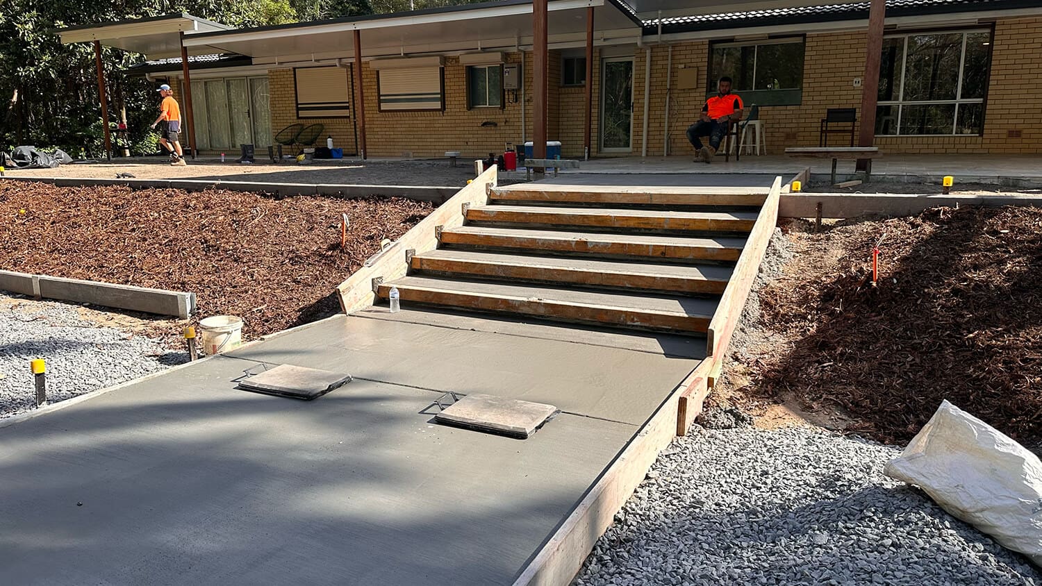 A freshly poured concrete ramp and steps in front of a single-story brick building, leading to the entrance. Workers are visible around the site, with one sitting on a chair near the door. The sides of the ramp are bordered by mulch and gravel, meticulously arranged by Pool Builders Brisbane.