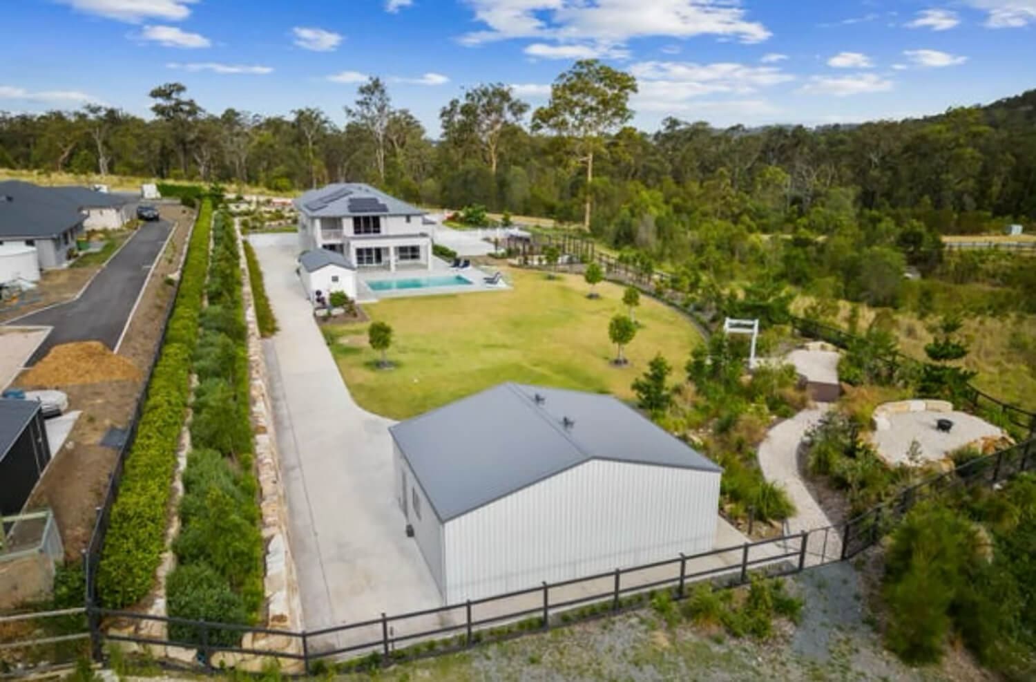 A large, modern home with a spacious backyard, pool, and patio sits on a lush, green property surrounded by trees. In the foreground, there is a long driveway leading to a metal-roofed outbuilding. The backyard includes various landscaped areas and a garden designed by top Pool Builders Brisbane.