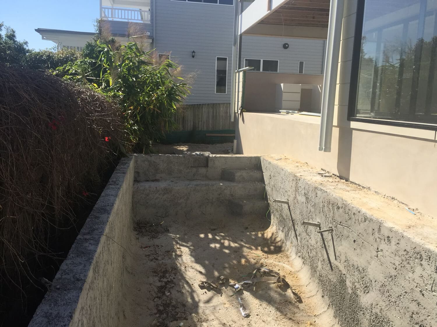 An empty rectangular concrete pool with steps sits in a backyard, hinting at the craftsmanship of Pool Builders Brisbane. Overgrown vegetation is seen on the left side, and the pool area appears unfinished or in need of repair. A modern house with large windows is adjacent to the pool area.