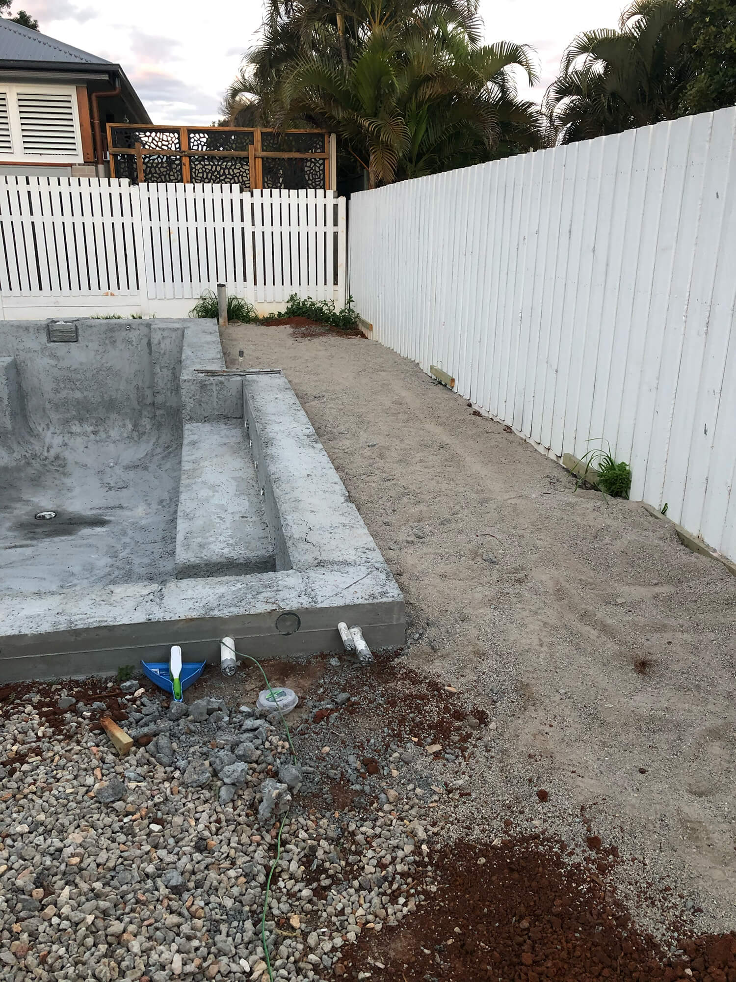 A backyard scene features an empty, unfinished swimming pool with exposed concrete, likely awaiting the expertise of Pool Builders Brisbane. The pool is surrounded by a gravel and sandy area. A white wooden fence encloses the space, while a section of garden with some plants is seen near the fence on the right.