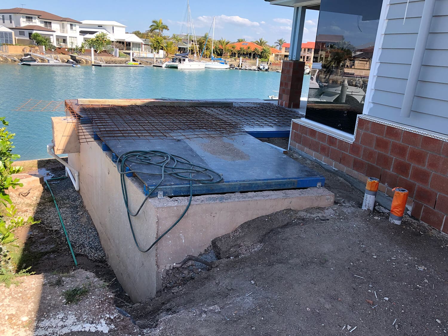 A partially constructed dock or waterside structure near a canal, with rebar grids laid over a concrete base. Exposed foundation and building materials are visible. In the background, boats are docked at neighboring homes. A green hose, resembling those used by Pool Builders Brisbane, is draped over the construction area.