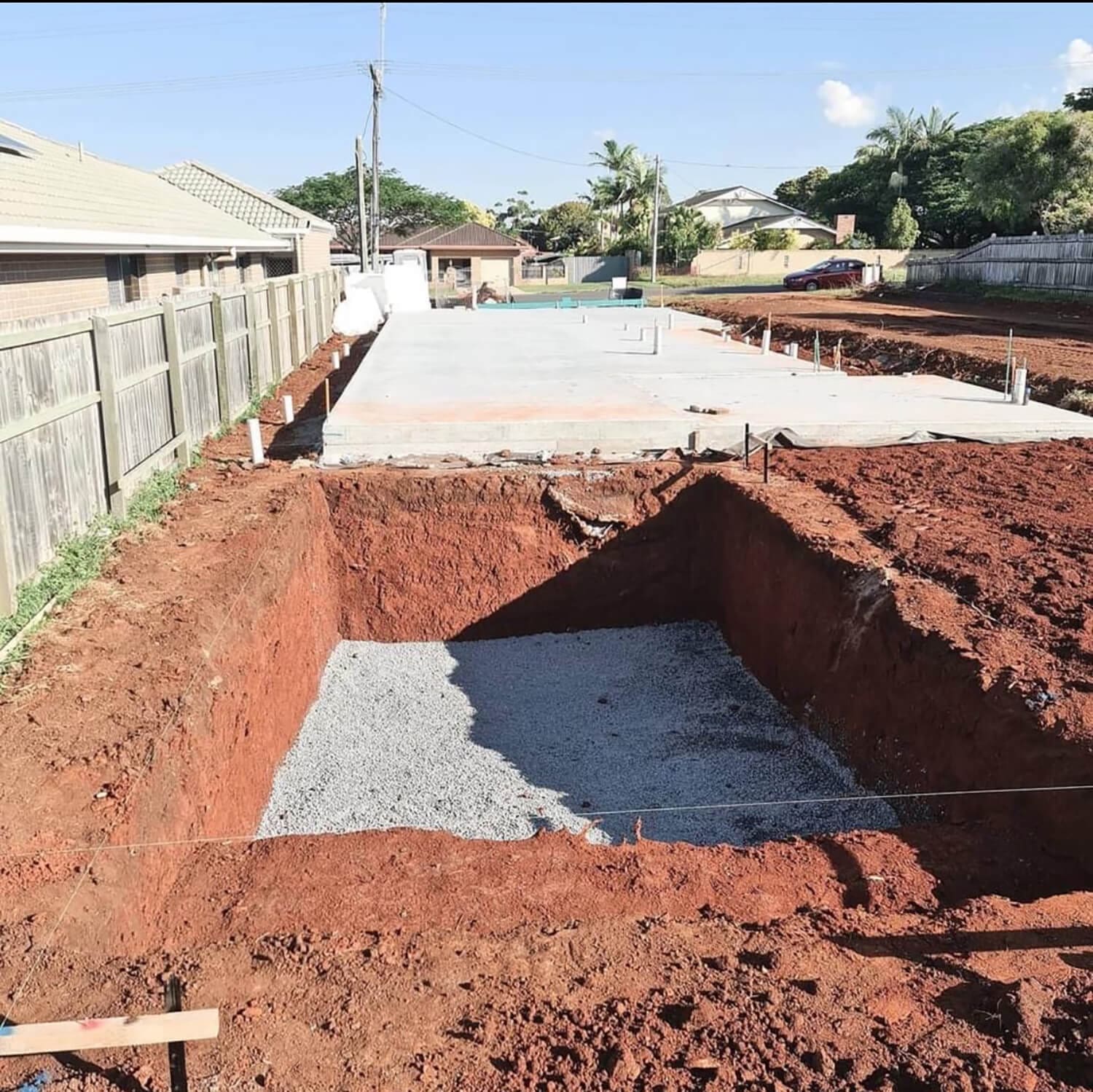 A construction site with an excavated hole filled with gravel in the foreground and a freshly laid concrete foundation in the background. The area, managed by Pool Builders Brisbane, is surrounded by a wooden fence on the left, houses, and power lines with a clear sky above.