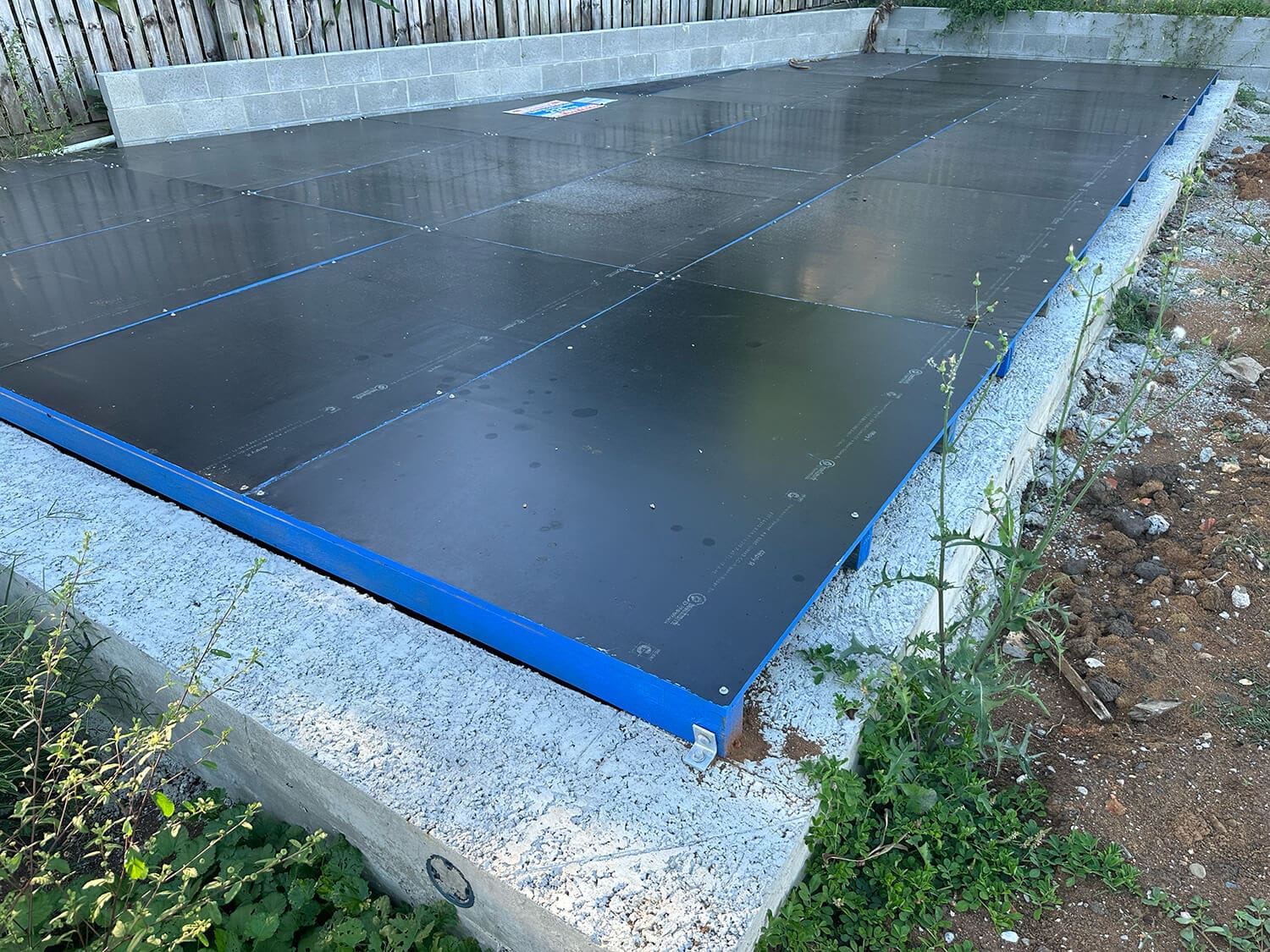 A construction site with the foundation of a building covered by black panels framed with blue edges. The concrete base extends around the covered area. Tall grass and weeds grow near the foundation, hinting at a future project possibly overseen by skilled pool builders in Brisbane. A wooden fence is visible in the background.