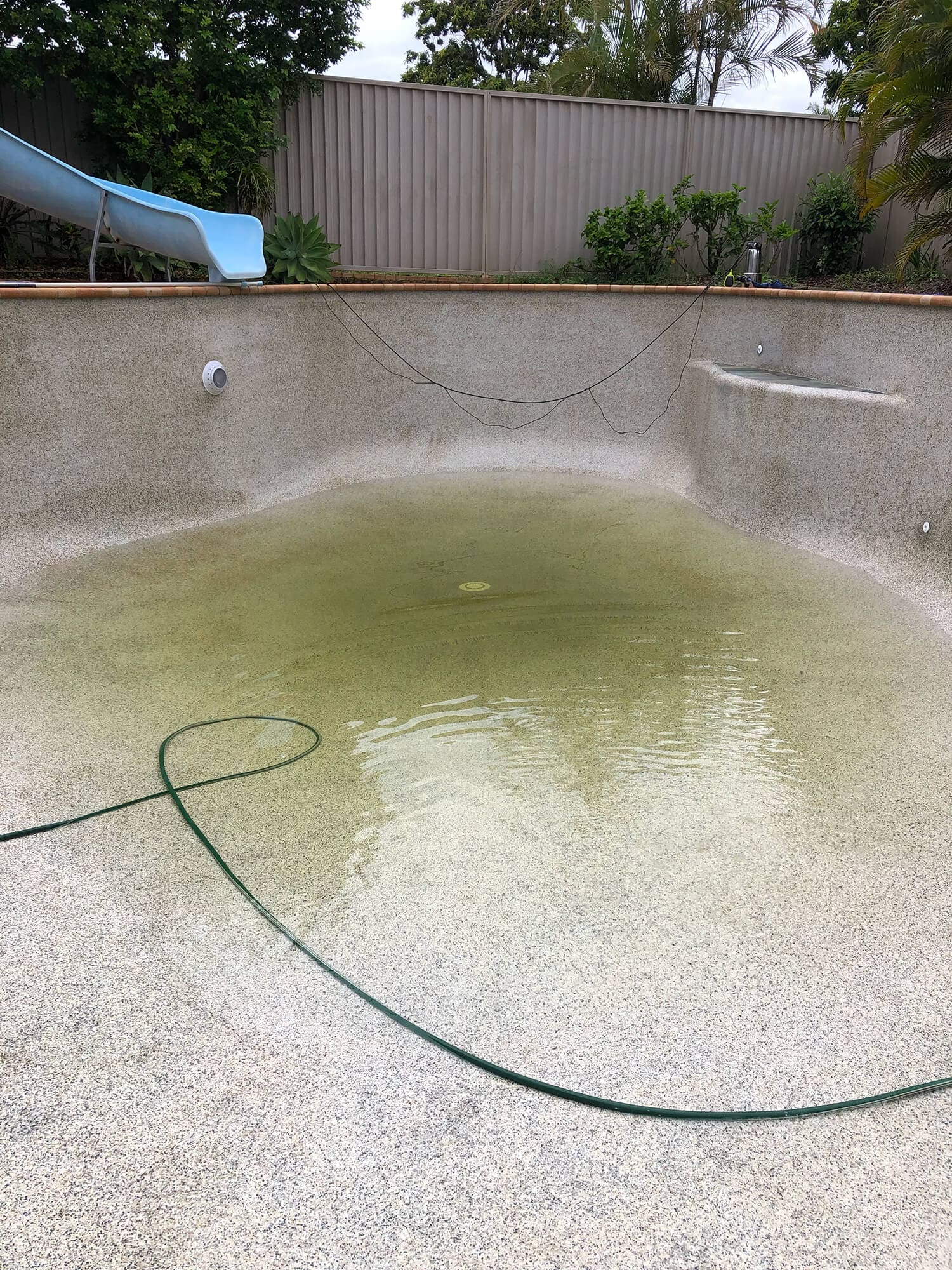 Partially filled swimming pool with dirty water at the bottom, surrounded by a tall wooden fence and some greenery. There is a blue slide on the left side and a garden hose lying on the pool floor—ideal for Pool Builders Brisbane to transform into an oasis.