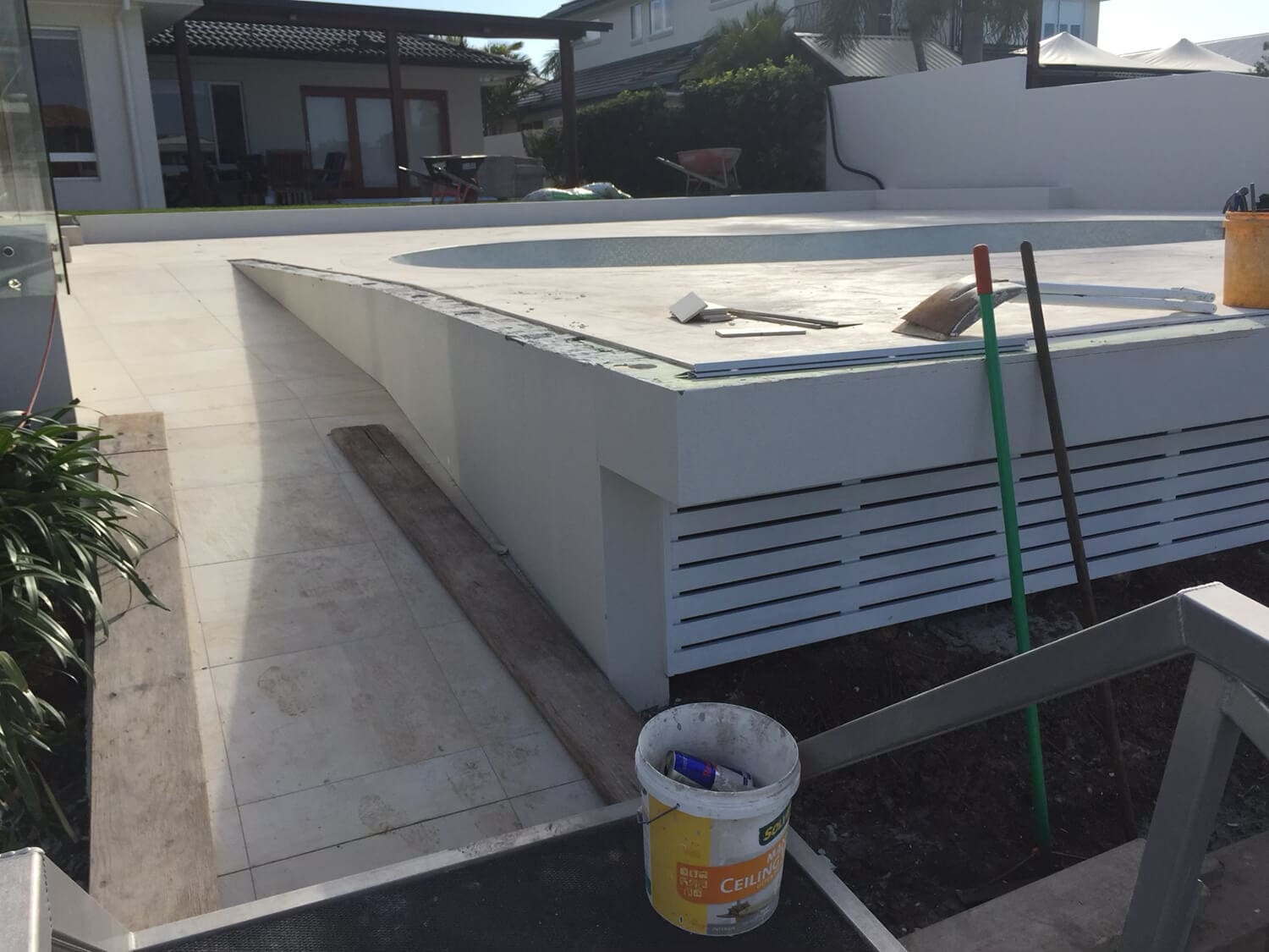 A construction site features an unfinished swimming pool with tools and construction materials scattered around. A bucket of cement sits in the foreground. A modern house with large windows is visible in the background, along with a wheelbarrow and some plants. Pool Builders Brisbane are on the job, ensuring quality work.