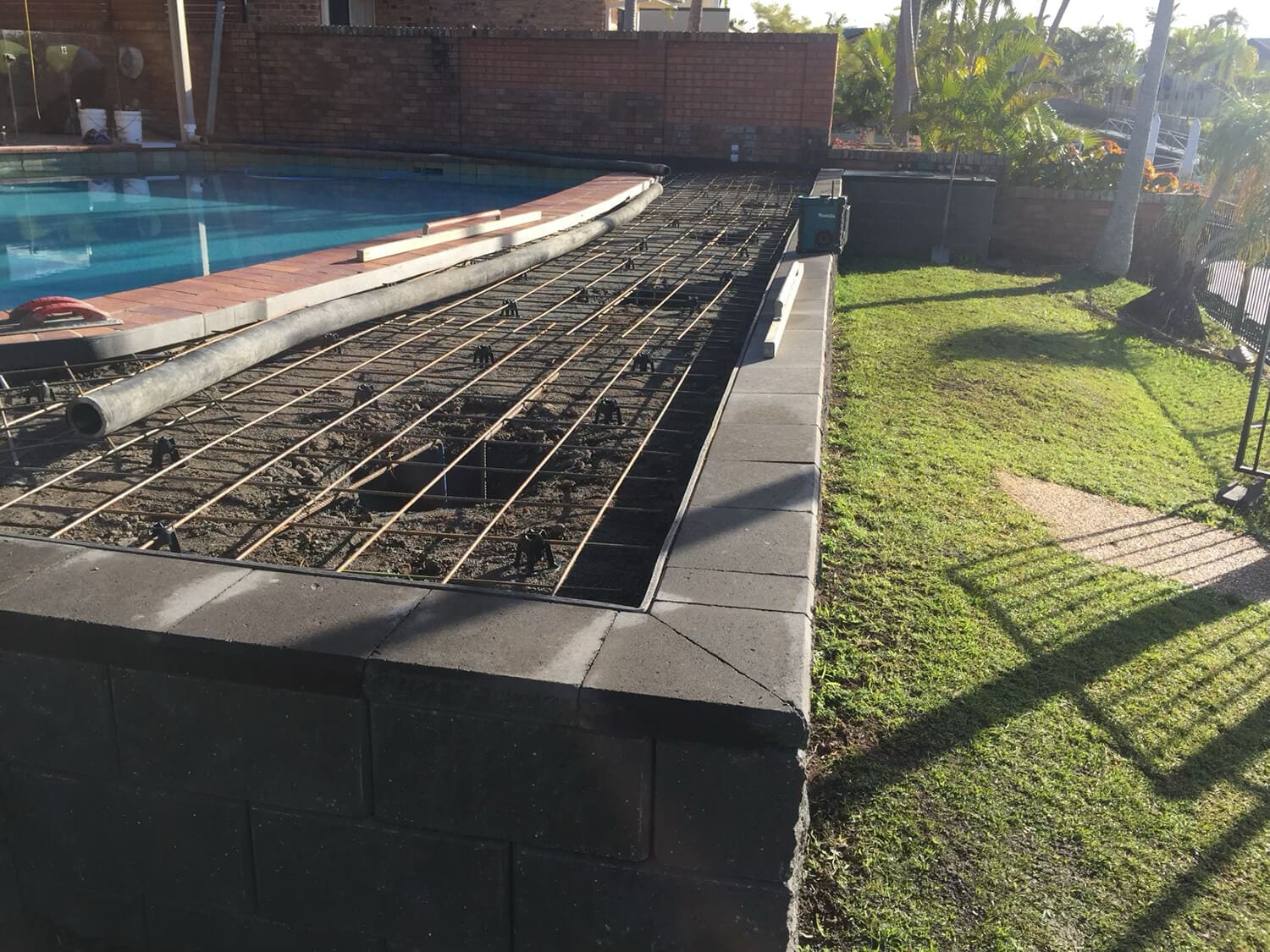 A swimming pool under construction with a metal rebar grid laid out beside it hints at the skilled work of Pool Builders Brisbane. Adjacent to the pool is a neatly mowed grassy area, with shadows of nearby objects and plants cast on the grass. A brick wall is visible in the background.