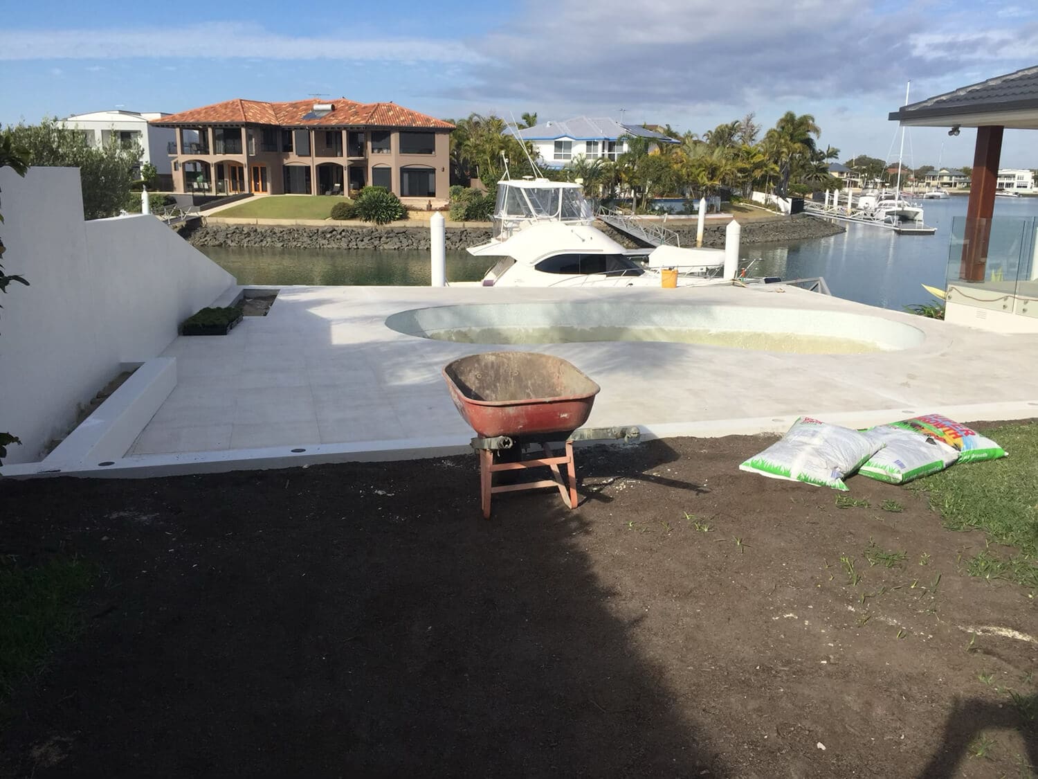 A construction site with a partially built pool near a waterfront. There's a red wheelbarrow in the foreground, bags of building materials, and luxurious houses, boats, and palm trees in the background under a partly cloudy sky. Pool Builders Brisbane are transforming this prime location into an oasis.