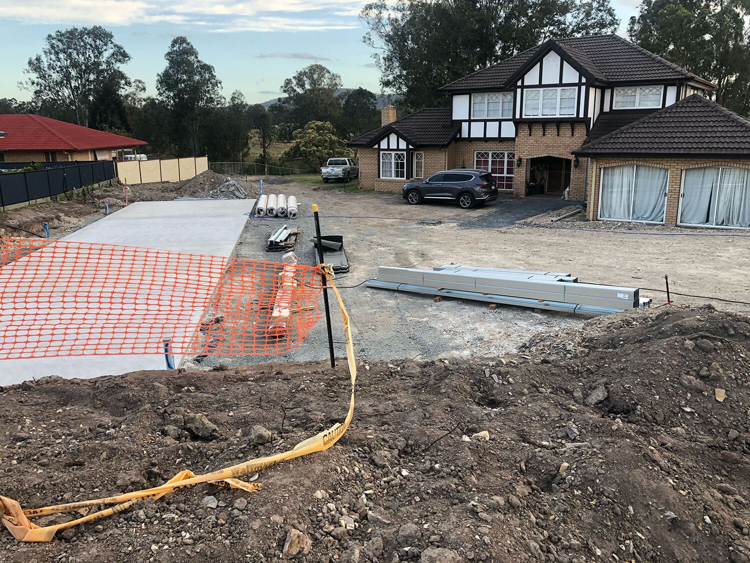 A construction site for a new driveway in front of a two-story house with Tudor-style architectural elements. The area, reminiscent of a work zone managed by Pool Builders Brisbane, is marked with orange safety fencing and scattered construction materials. A silver car is parked nearby, trees visible in the background.