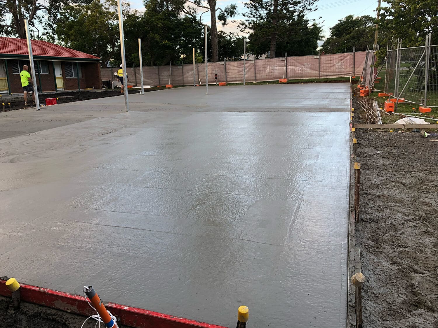 A freshly poured concrete slab at a construction site, bordered by wooden framing and surrounded by dirt. Several workers, possibly from Pool Builders Brisbane, are visible in the background near a fence and a building with a red roof. Trees and greenery are also present in the background.