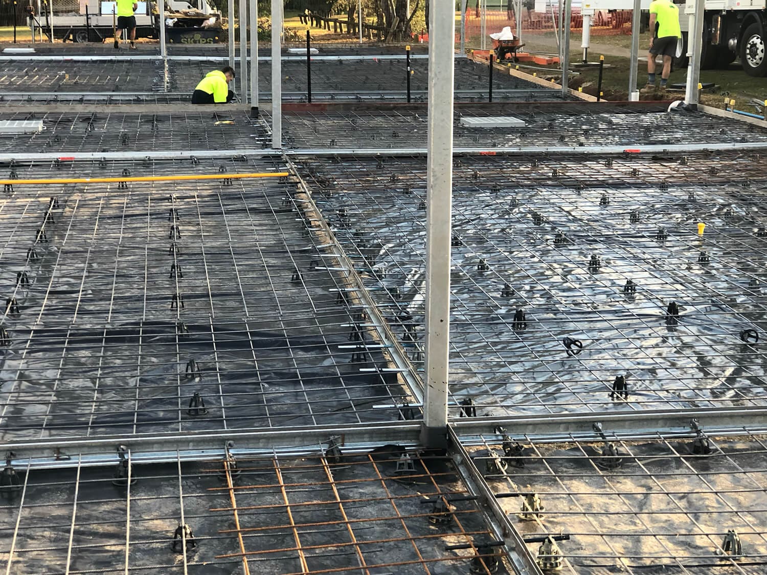 Construction site with multiple concrete foundations prepped with rebar and plastic sheeting. Workers in yellow vests and hard hats are seen in the background working on various tasks. Metal poles are installed around the site, indicative of a building framework, much like projects by Pool Builders Brisbane.