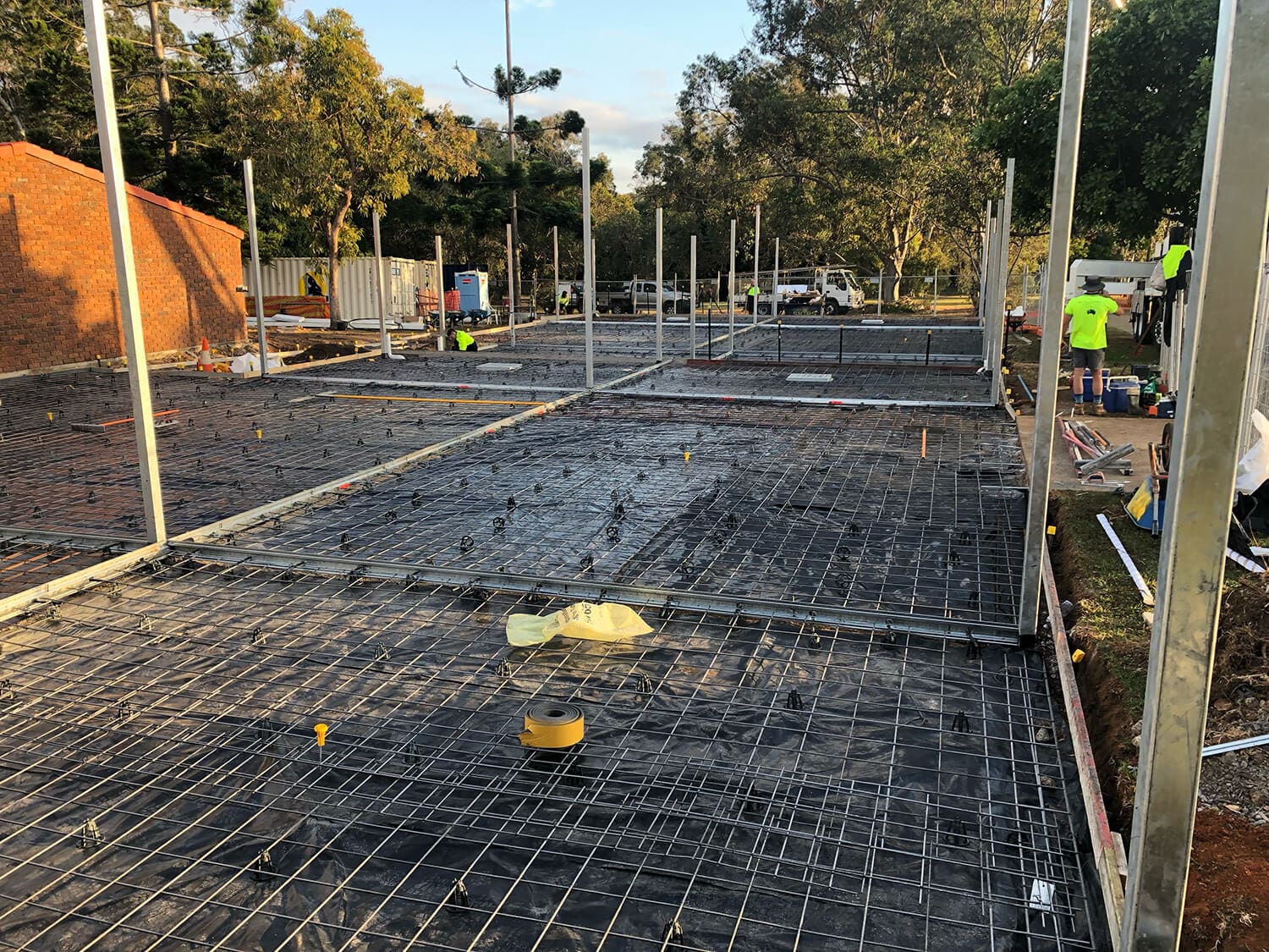 Construction site with metal framing and rebar grid laid on the ground for a building foundation. Several workers in high-visibility vests are present, with some equipment and materials scattered around. Trees and a brick structure are visible in the background, suggesting Pool Builders Brisbane at work.