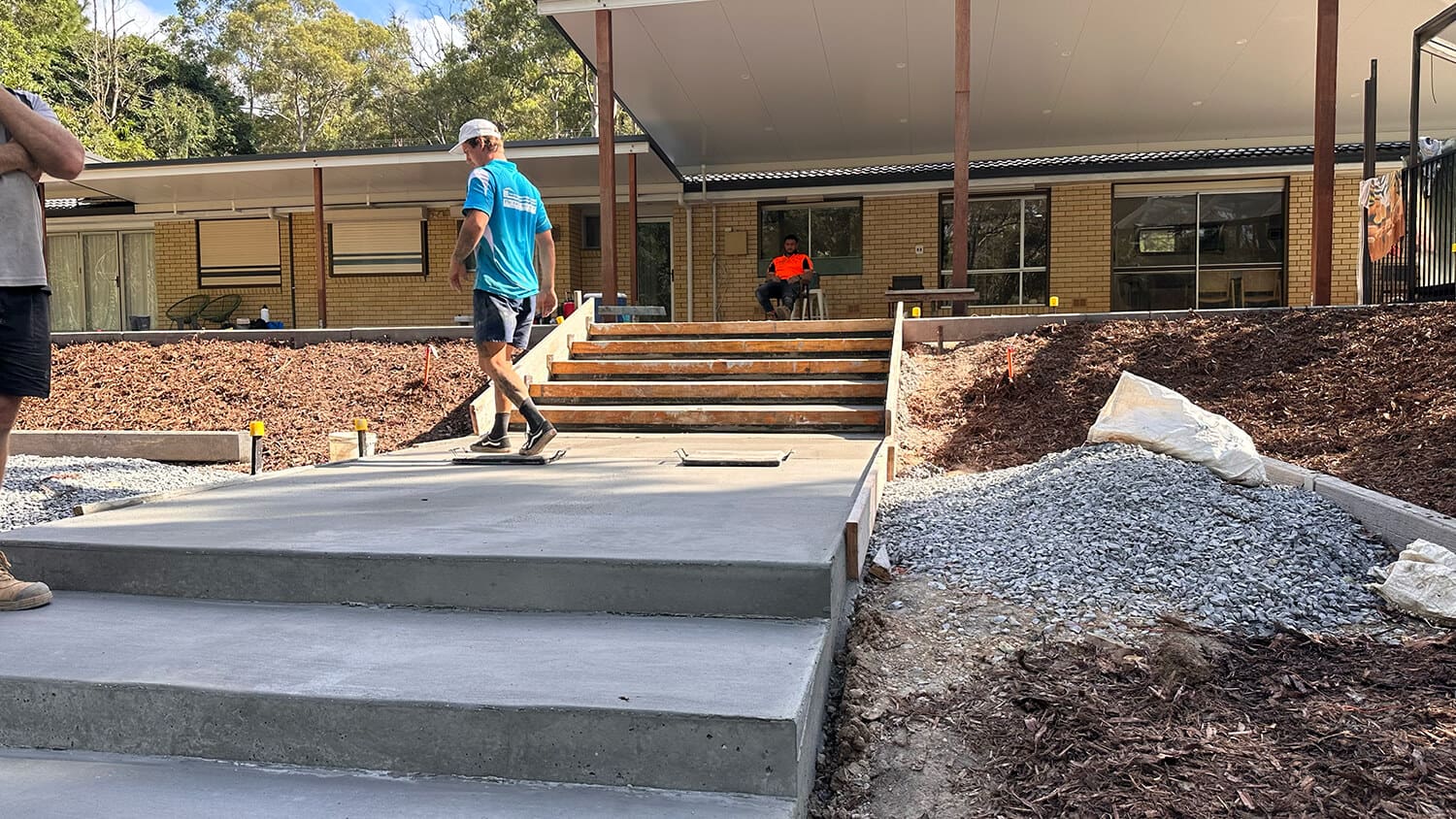 A construction worker in a blue shirt and white hard hat walks on a newly built concrete path with steps leading to a building. Another worker in an orange shirt, perhaps planning his next project with Pool Builders Brisbane, sits on the steps. The surroundings include mulch and gravel. Trees and a clear sky are visible in the background.