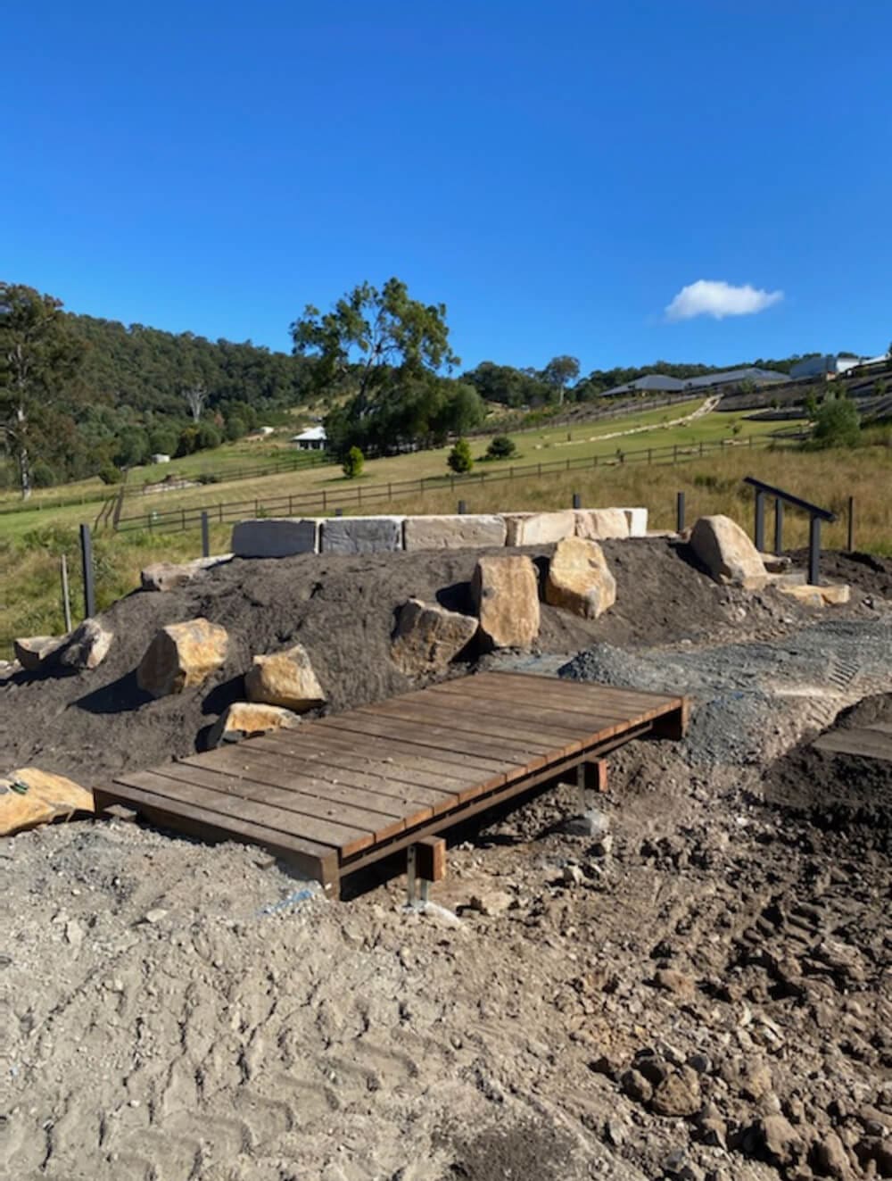 A wooden bridge crosses a dirt and rock path in an outdoor environment curated by Pool Builders Brisbane. Large rocks are scattered around, and grassy hills with scattered trees can be seen in the background under a clear blue sky.