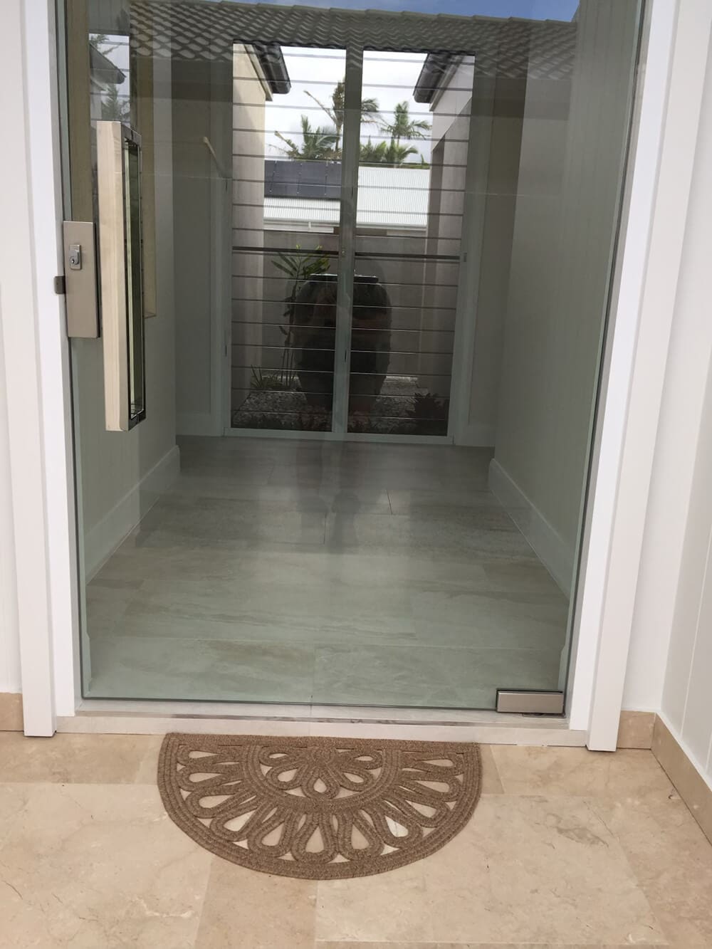 A modern entrance with a glass door, framed by white walls, opens to a narrow hallway with a large pot plant at the end. A half-circle doormat with a decorative pattern rests atop light-colored floor tiles. Reflective surfaces and clean lines emphasize minimalistic design, much like the work of top Pool Builders Brisbane.