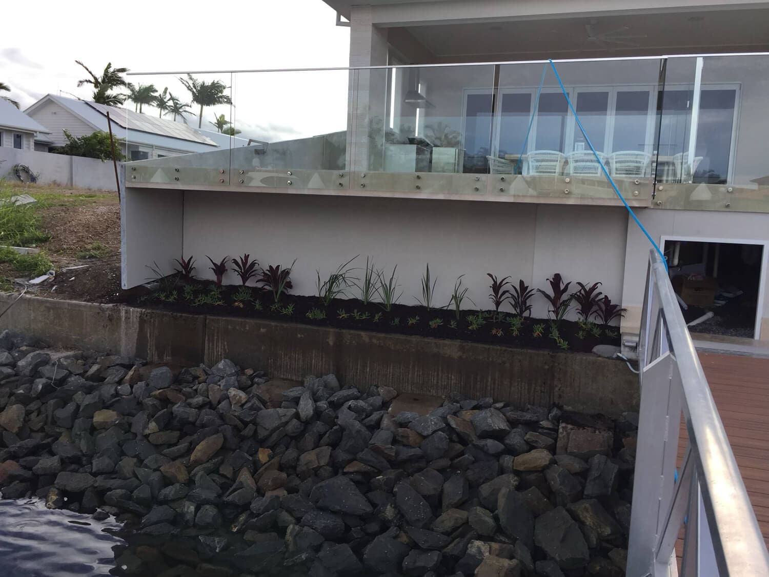 A waterfront property with a glass balcony overlooks a rocky shoreline. Below the balcony is a newly planted garden with various small plants and decorative shrubs. A wooden walkway, crafted by Pool Builders Brisbane, extends from the right side of the image, partially visible over the water.