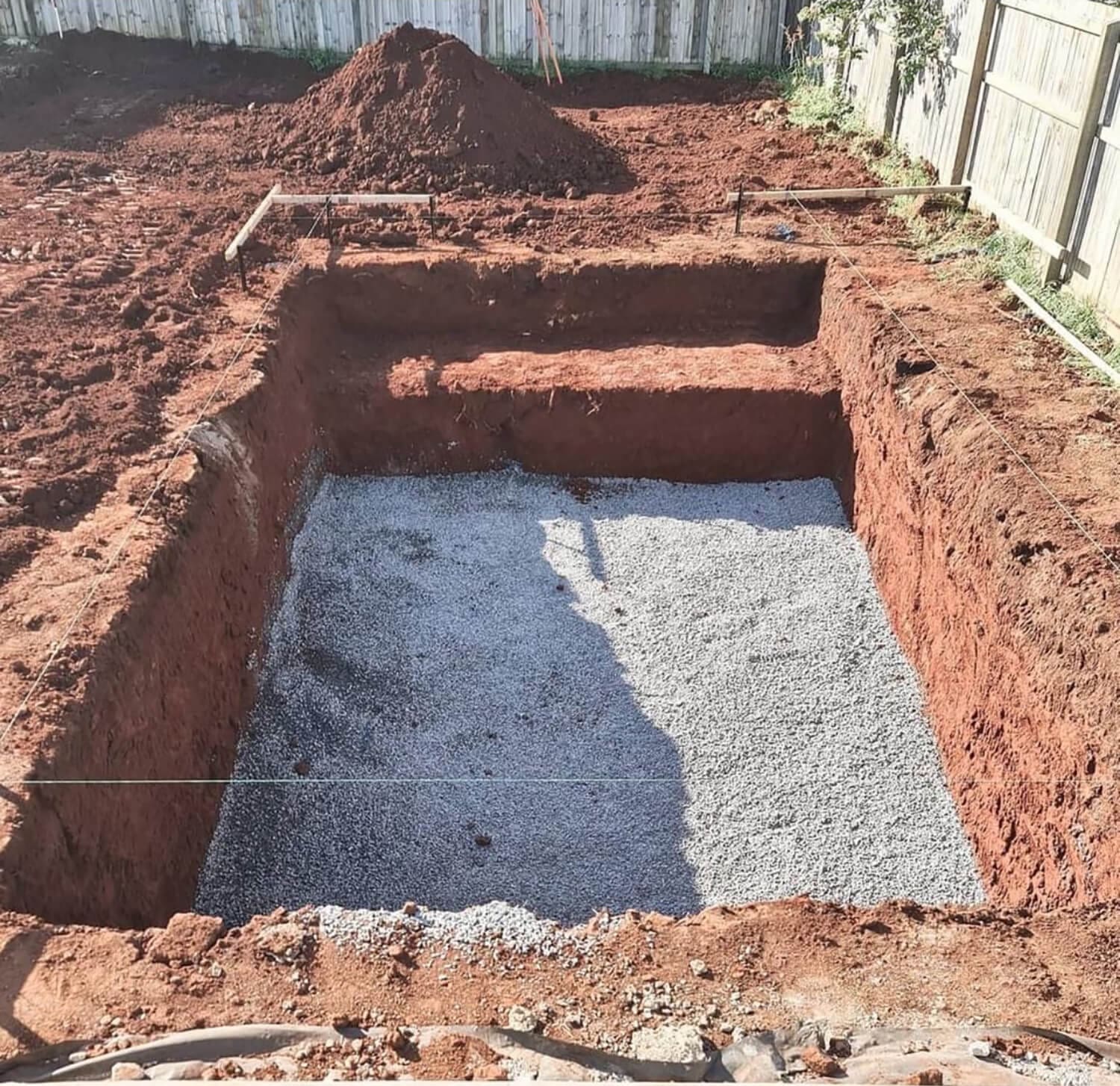 A rectangular excavation site with reddish soil, featuring two levels, is shown. The lower level is lined with a layer of gravel, while the upper level remains just soil. The background has a wooden fence and a pile of dug-up soil, common in projects by Pool Builders Brisbane. String lines outline the excavation.