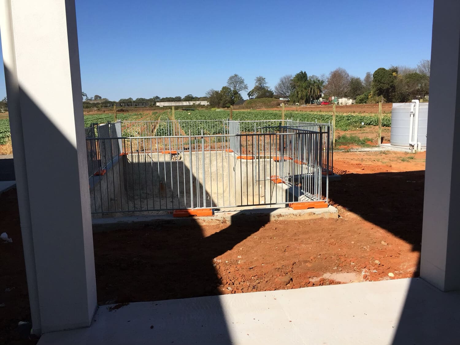 A concrete slab is enclosed by a metal fence with an orange safety barrier at the corners, likely set up by pool builders in Brisbane. It is surrounded by dirt ground that extends to fields of crops in the background. The scene is viewed from a shaded concrete structure in the foreground.