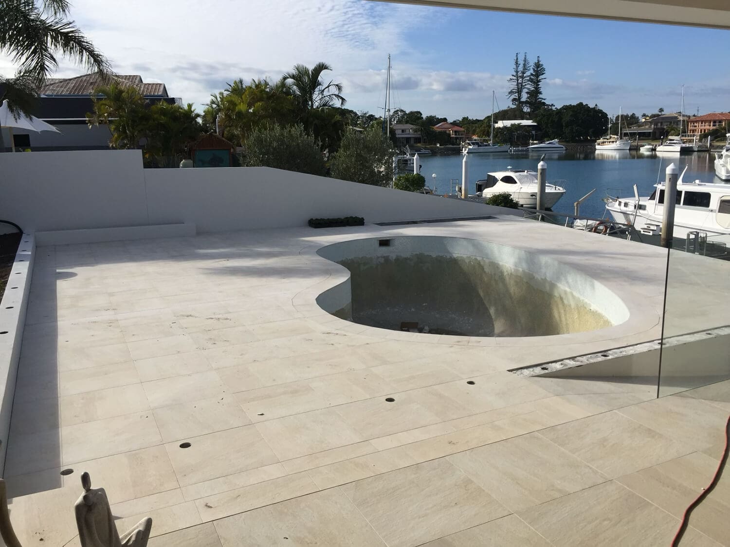 A backyard pool area under construction, overlooking a marina with several anchored boats. The pool itself is empty and appears to be in the midst of renovation by Pool Builders Brisbane. The surroundings feature beige tile flooring and a partial glass fence. Palm trees and houses are visible in the background.