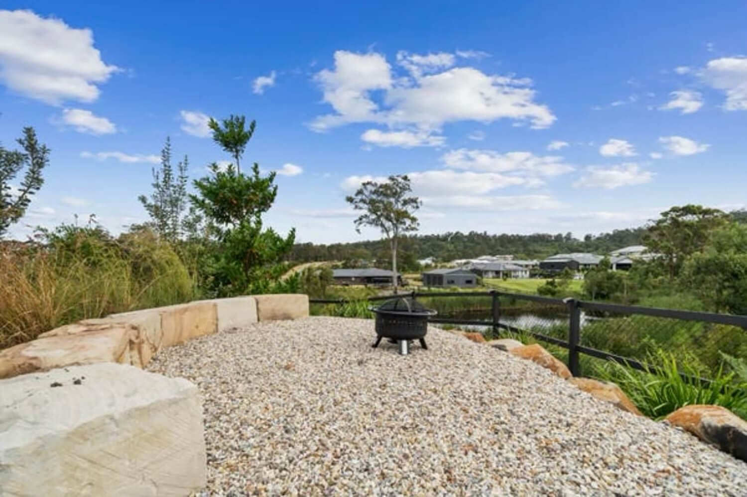 A peaceful outdoor space with a gravel ground featuring a small fire pit in the center. There are large stone blocks used as seating around the area. The backdrop includes green trees, shrubs, and a scenic view of nearby houses under a blue sky, perfect for Pool Builders Brisbane to draw inspiration from.