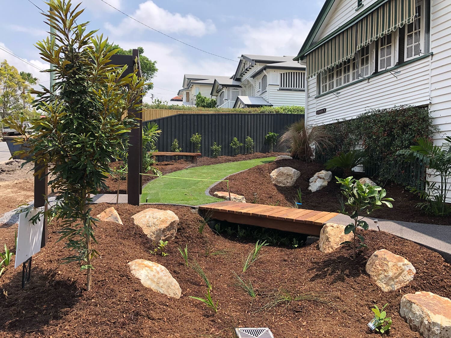 A landscaped garden with a curved pathway, wooden bridge, and large rocks surrounded by mulch. Newly planted shrubs and trees add charm. A white house with green accents and a striped awning is on the right. Benches line the fence in the background, enhancing the serene setting ideal for Pool Builders Brisbane to create your dream pool oasis.