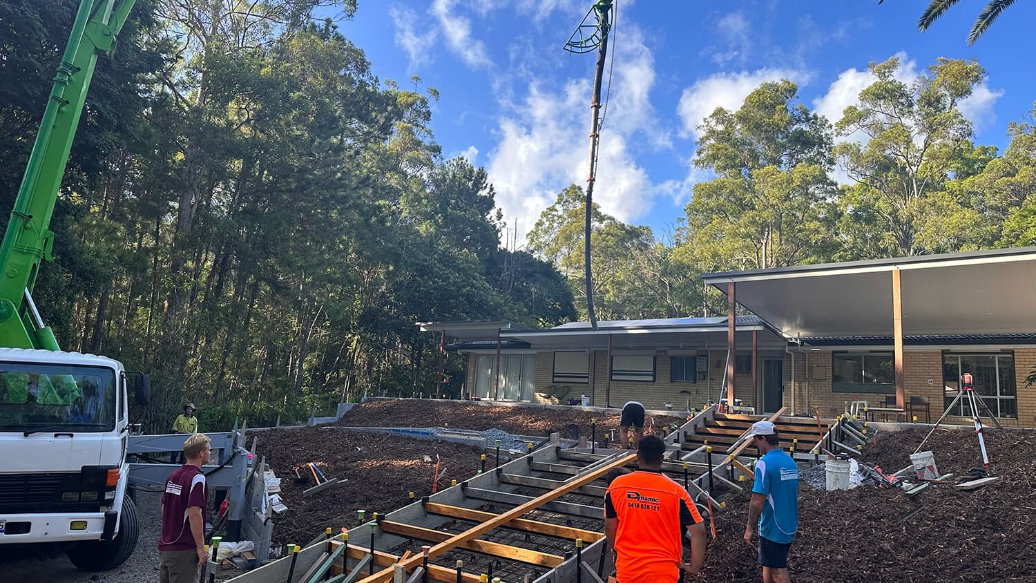 A group of workers at a construction site are installing wooden framework for stairs outside a building surrounded by trees. A crane and truck from Pool Builders Brisbane are on site. The building is modern with a large covered patio on the right. The sky is clear with scattered clouds.