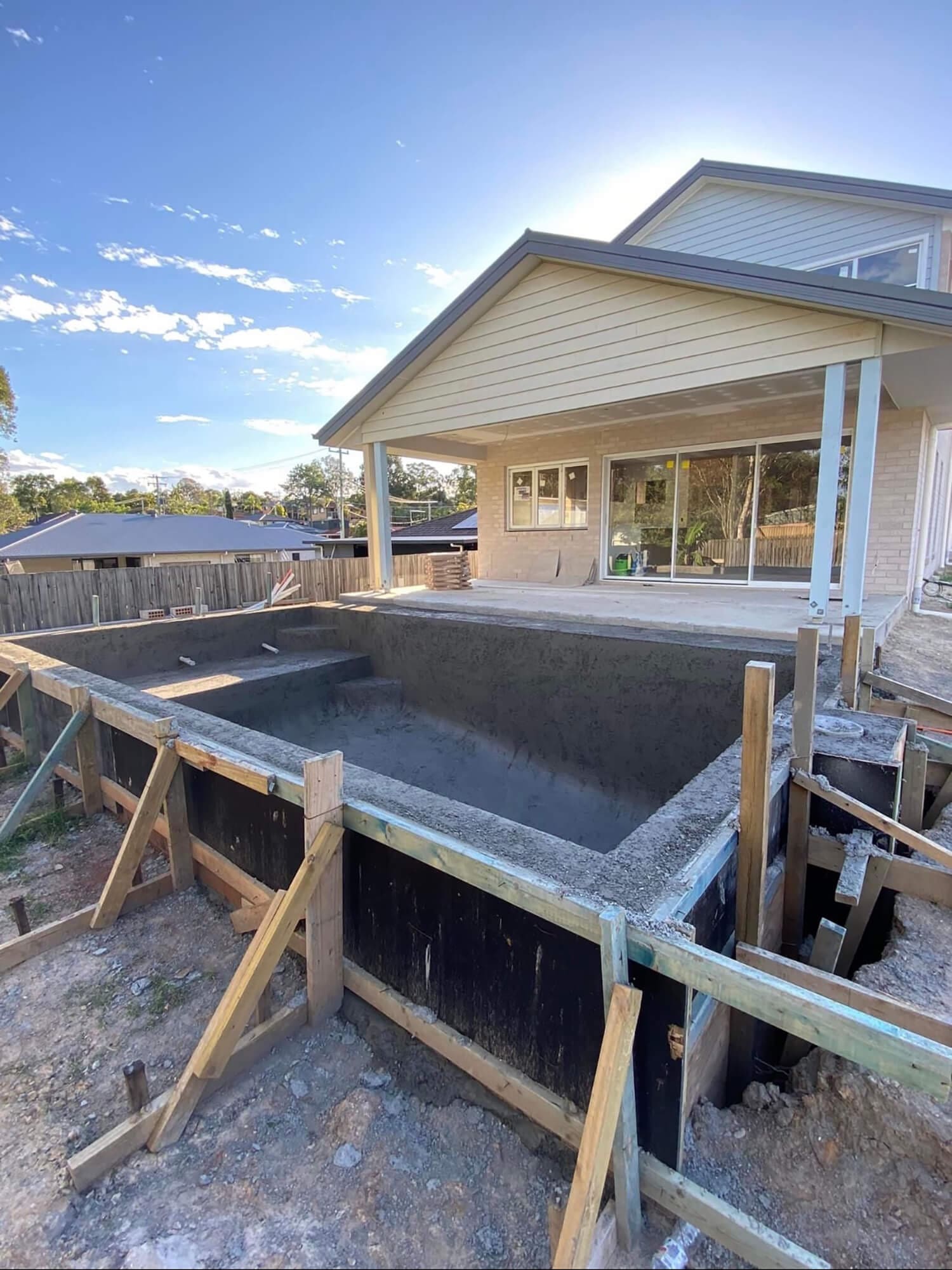 An in-ground swimming pool by Adella Pools under construction in a backyard. Wooden framing supports the concrete structure, and the pool is adjacent to a house with large windows and a covered patio. The sky is clear and sunny—perfect conditions for Pool Builders Brisbane to work their magic.