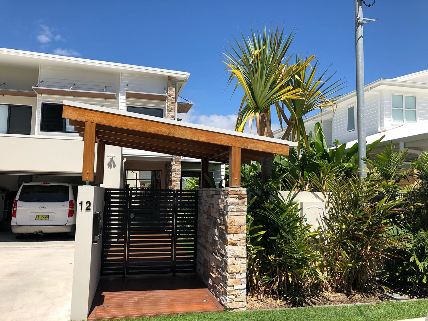 A modern house with a double garage and a white car parked in the driveway. The entrance has a wooden gate with stone pillars and a house number "12" displayed. Lush greenery surrounds the area, with Adella Pools by Pool Builders Brisbane adding charm. Two similar white houses are adjacent under a clear blue sky.