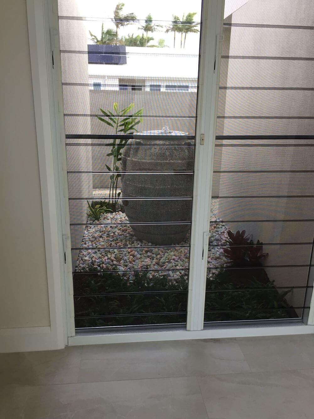 View of an outdoor area through a large sliding glass door. The courtyard features a tall, large stone water feature surrounded by small rocks and plants. Expertly crafted by Adella Pools, the outdoor space is enclosed by high concrete walls, with a glimpse of the sky and palm trees above.