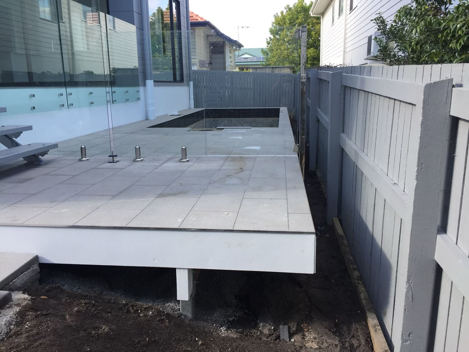 A concrete patio with glass railings under construction next to a gray wooden fence. The patio is raised, with visible support beams underneath. A house with large windows sits on one side, and another building and greenery provide a picturesque backdrop—courtesy of Adella Pools, renowned pool builders Brisbane trusts.