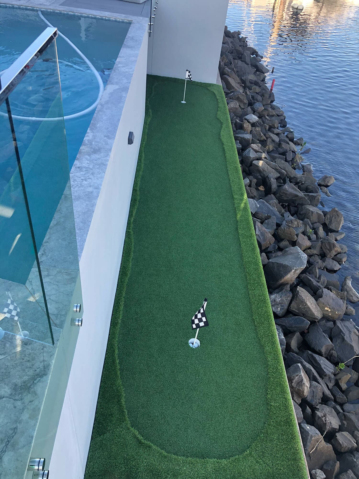 A narrow mini-golf green runs alongside a body of water with a glass barrier separating it from a swimming pool on the left, masterfully crafted by Adella Pools. A checkered flag is placed in the hole on the green. Rocks and stones line the edge of the water on the right.