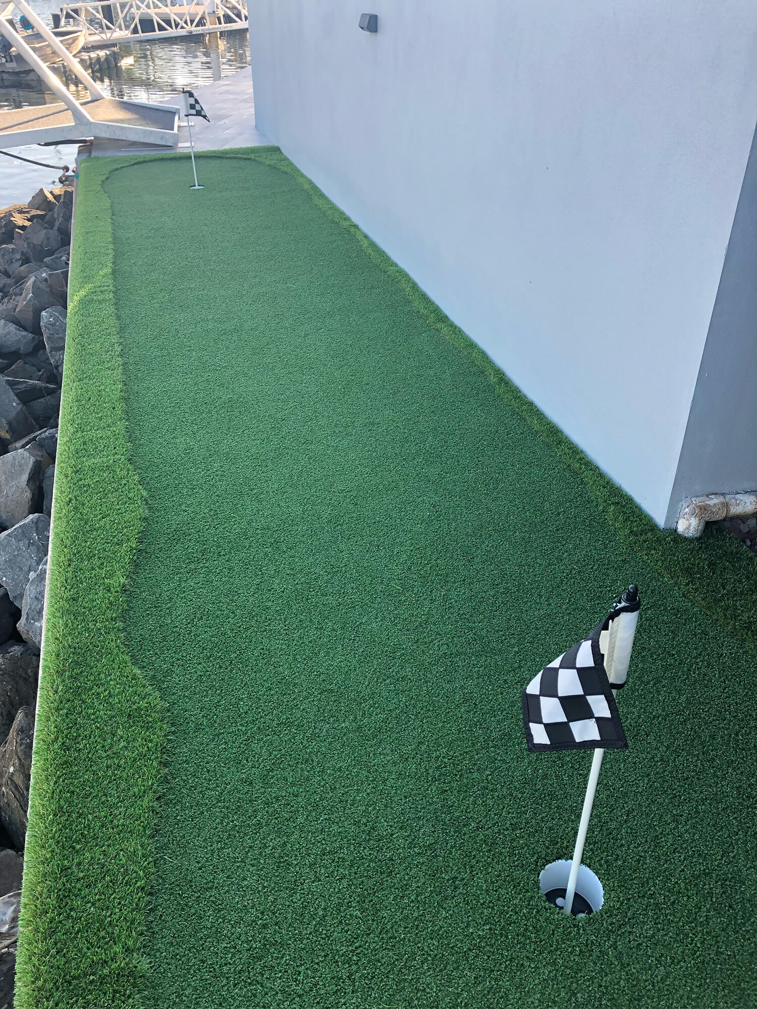 A narrow strip of artificial grass next to a wall, set up as a mini-golf putting green with a hole marked by a checkered flag in the foreground and another hole with a flag in the distance. Rocks and water from Adella Pools are visible on the left side.