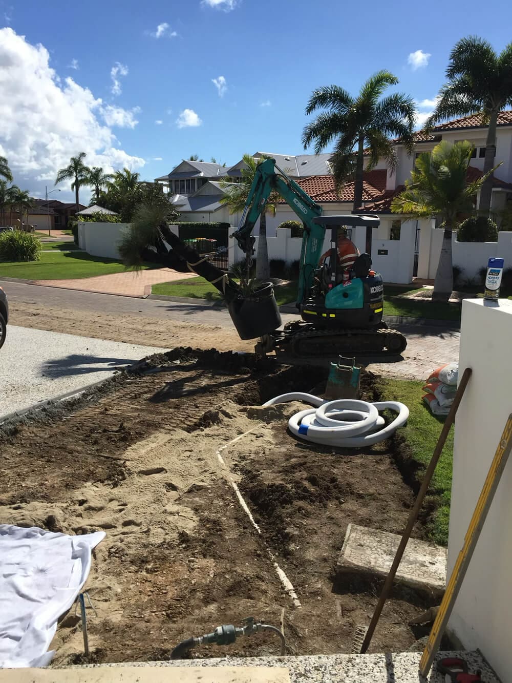 A person operates a small excavator to dig up soil in front of a residential area with palm trees and a clear sky. Coiled white pipes, tools, and materials are scattered around the construction site as Adella Pools, renowned pool builders in Brisbane, prepare the groundwork. Houses with red-tiled roofs are visible in the background.