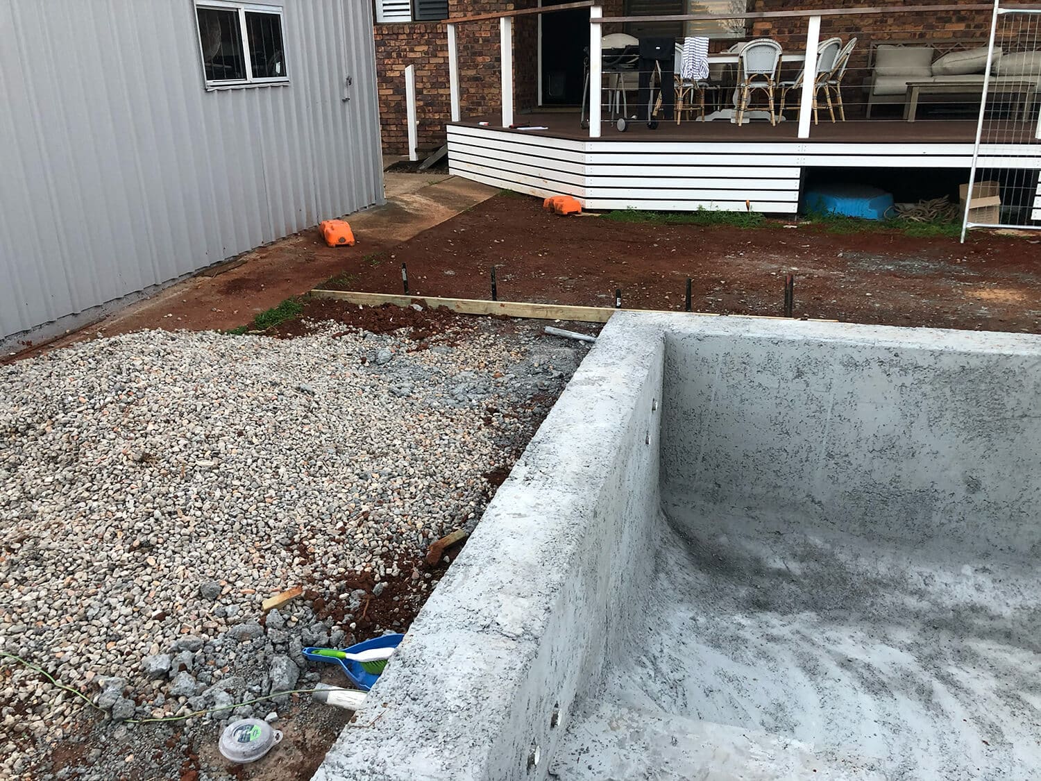 A partially constructed pool with a concrete shell by Adella Pools sits next to a pile of gravel in a backyard. A wooden deck with outdoor furniture is visible in the background, attached to a brick house. Orange construction barriers and various tools from Pool Builders Brisbane are scattered around.