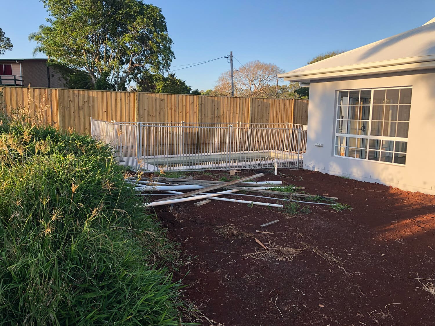 A fenced garden area beside a white house, with a pile of metal pipes and wooden debris on the ground in a dirt area. Tall grass grows in the foreground, and a wooden fence lines the background. A large tree and clear sky are visible, making it an ideal spot for Adella Pools to work their magic as top Pool Builders Brisbane trusts.