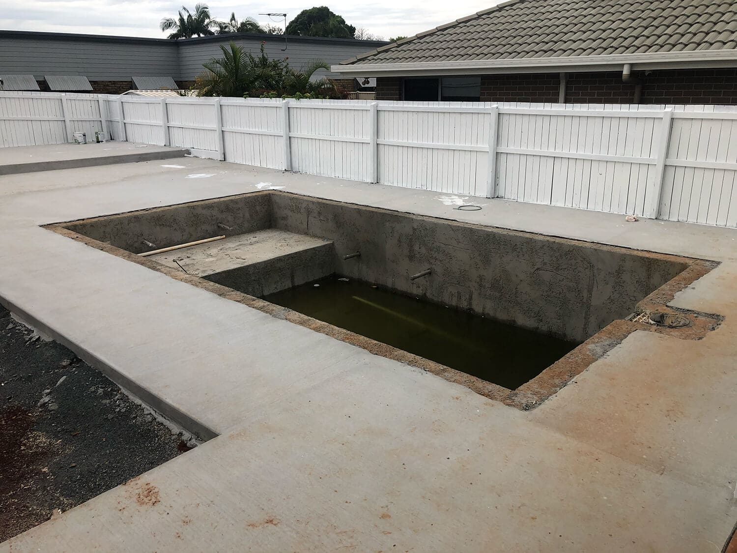 An empty, rectangular swimming pool under construction by Adella Pools is set in a concrete backyard. The pool is partially filled with murky water at the bottom. White picket fences surround the area, and a house with a tiled roof is visible in the background.