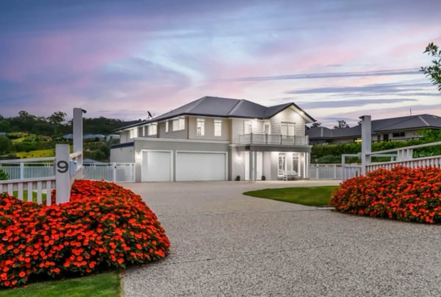 A two-story modern house with a grey roof and exterior, large garage door, and balcony is seen framed by a vibrant garden with red flowers. A paved driveway leads to the house, surrounded by a white picket fence. Adella Pools recently added an elegant swimming pool to this picturesque home against a colorful twilight sky.