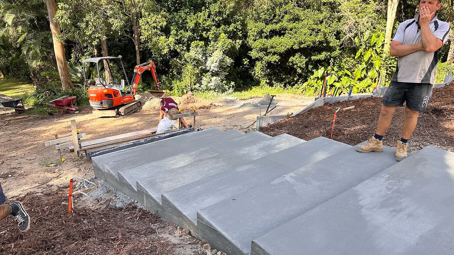 A person in workwear stands at the top of newly constructed concrete steps, with another working near the bottom and an excavator in the background. The area, surrounded by greenery and construction materials, is a busy site for Pool Builders Brisbane from Adella Pools.